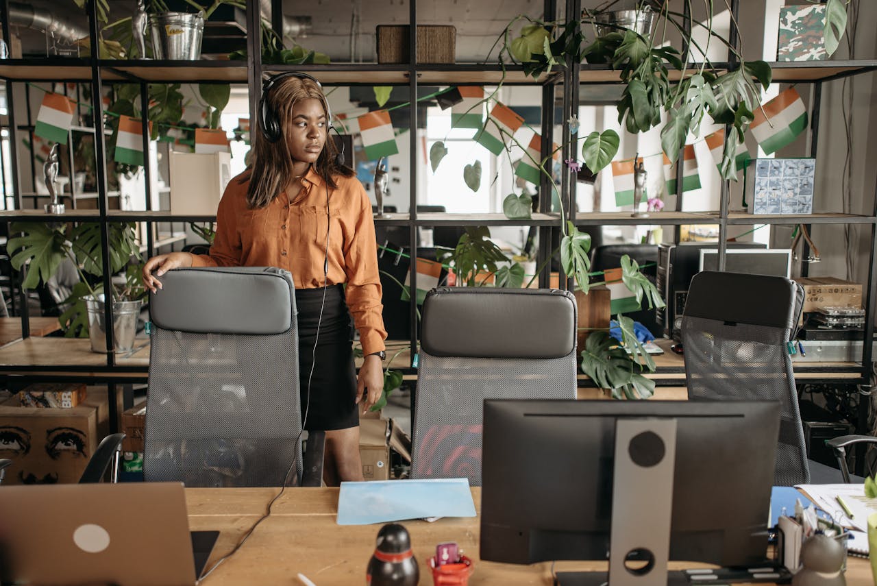 Businesswoman in orange shirt in modern office setting with plants and flags.